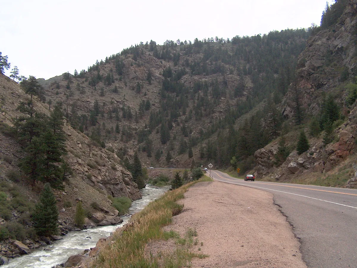 Clear Creek runs through a steep, rocky canyon beside U.S. Route 6, with pine-covered walls rising sharply on both sides. The morning light is soft and overcast, and the creek churns past boulders along the road’s edge.