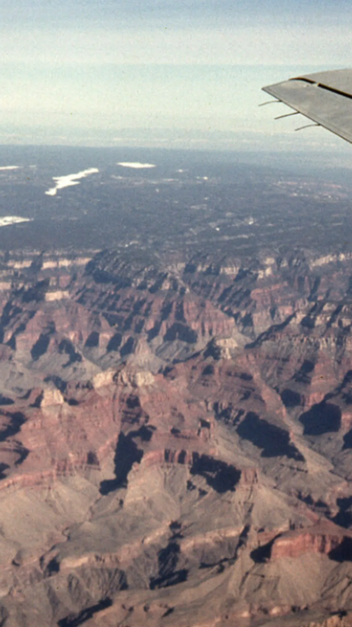 An aerial view looks down over the vast, layered red-and-brown walls of the Grand Canyon, with deep shadows carving out ridges and buttes under a hazy sky. The airplane wing enters from the upper right, adding a sense of height and motion above the rugged landscape.