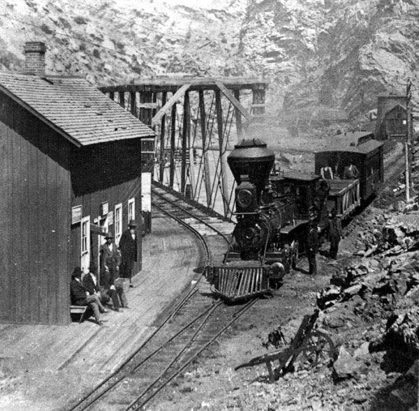 A narrow-gauge steam locomotive sits on curved tracks in a steep rocky canyon, with a steel truss bridge spanning the creek behind it. Wooden buildings hug the left side of the tracks, while a few people stand and sit beside the depot in the morning light.