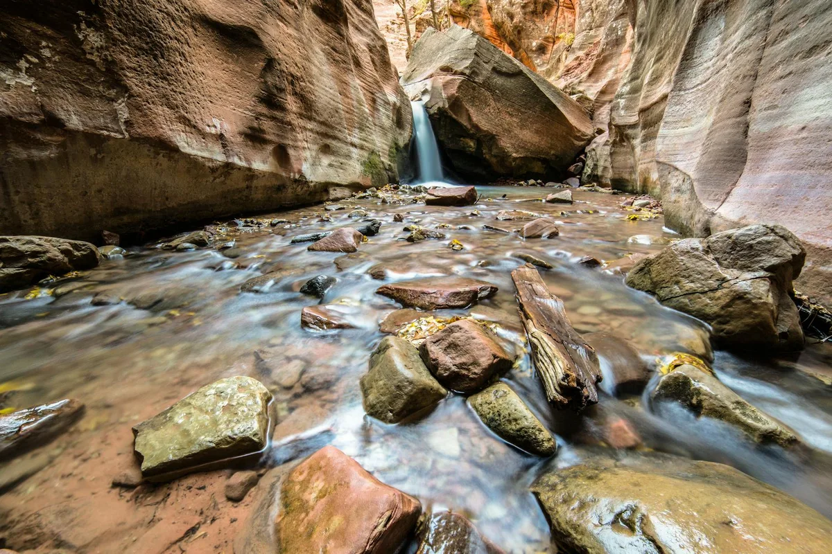 A narrow sandstone canyon frames a silky long-exposure creek flowing around scattered boulders and a weathered log. Soft morning light brightens the steep rock walls, while a small waterfall drops from beneath a tilted slab in the center background.