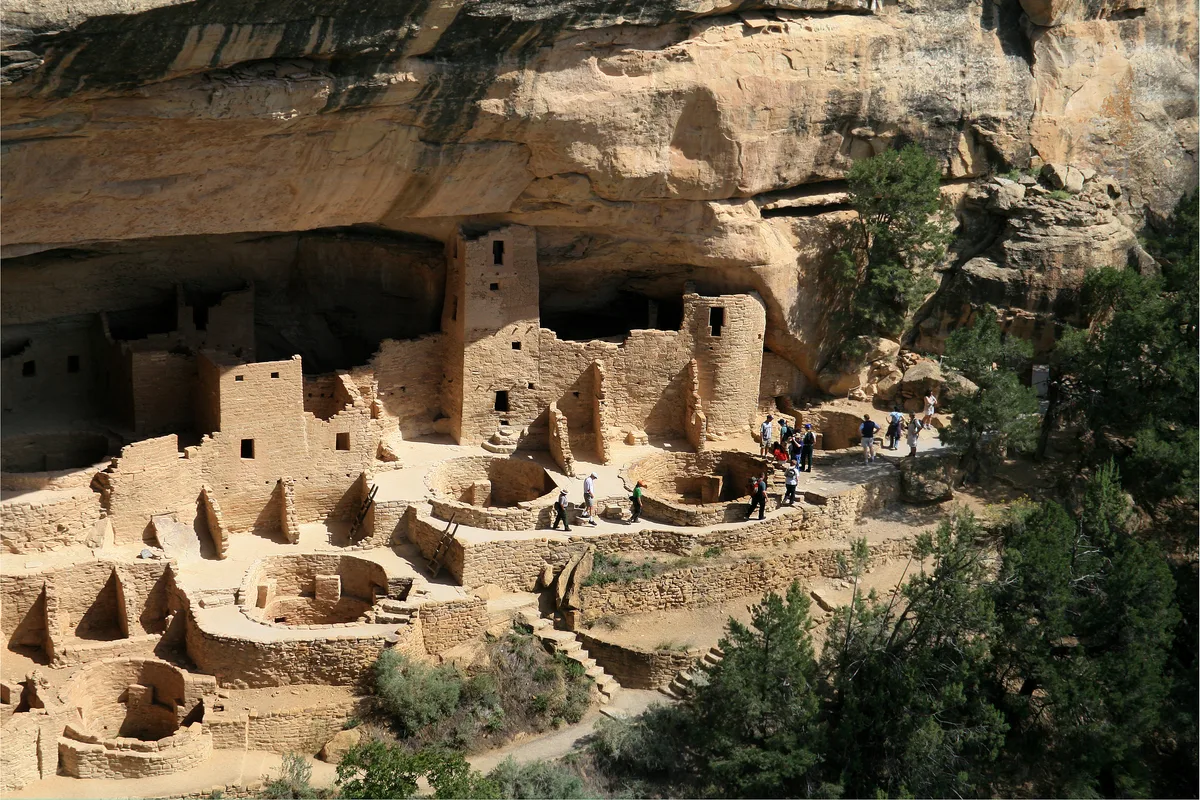 Sunlit sandstone ruins of Cliff Palace fill the alcove beneath a massive rock overhang, with round kivas, layered stone walls, and narrow towers clustered along the canyon ledge. Small groups of visitors stand on the pathways, while dark shade inside the cliff contrasts with the bright afternoon light on the weathered masonry.