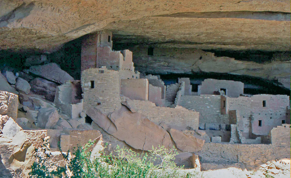 Sunlit stone rooms and window openings of Cliff Palace fill the alcove beneath a massive sandstone overhang, with layered canyon rock and scattered boulders framing the ancient cliff dwelling. Soft afternoon light brings out the warm tan and gray textures of the masonry against the shadowed cave ceiling.