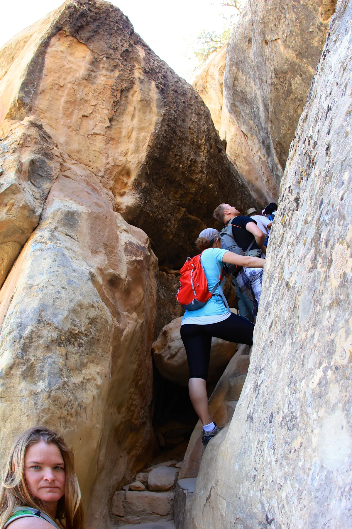 Visitors climb a narrow stone passage between towering sunlit sandstone walls at Cliff Palace, with one woman in a red backpack stepping up the rocks while another looks back from the lower left. The tight alcove is shaded in the center, emphasizing the scale and rough texture of the cliff dwelling’s canyon setting.