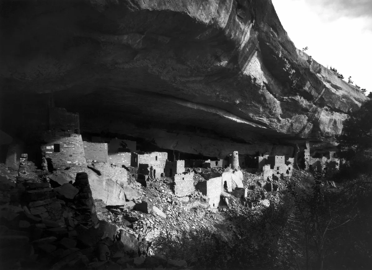 Wide black-and-white view of Cliff Palace tucked beneath a massive sandstone overhang, with rows of ancient stone rooms and kivas built into the shaded alcove. Sunlight highlights the front walls and rubble-strewn canyon floor, while the cliff ceiling looms dark overhead.