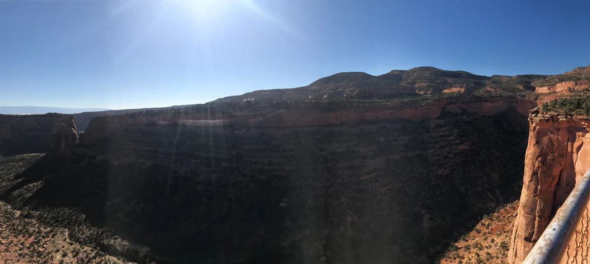 Bright afternoon sun floods a vast red-rock canyon, with steep shadowed walls dropping away below a rim lined by rust-colored cliffs and sparse desert vegetation. A metal railing at the right edge emphasizes the dizzying overlook across Columbus Canyon beneath a clear blue sky.
