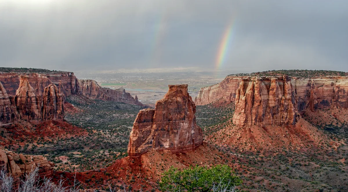 A sweeping view over red sandstone cliffs and deep canyons in Colorado National Monument, with a tall freestanding rock formation centered in the foreground. Dark storm clouds hang overhead while a faint rainbow arcs in the distance above the hazy valley.