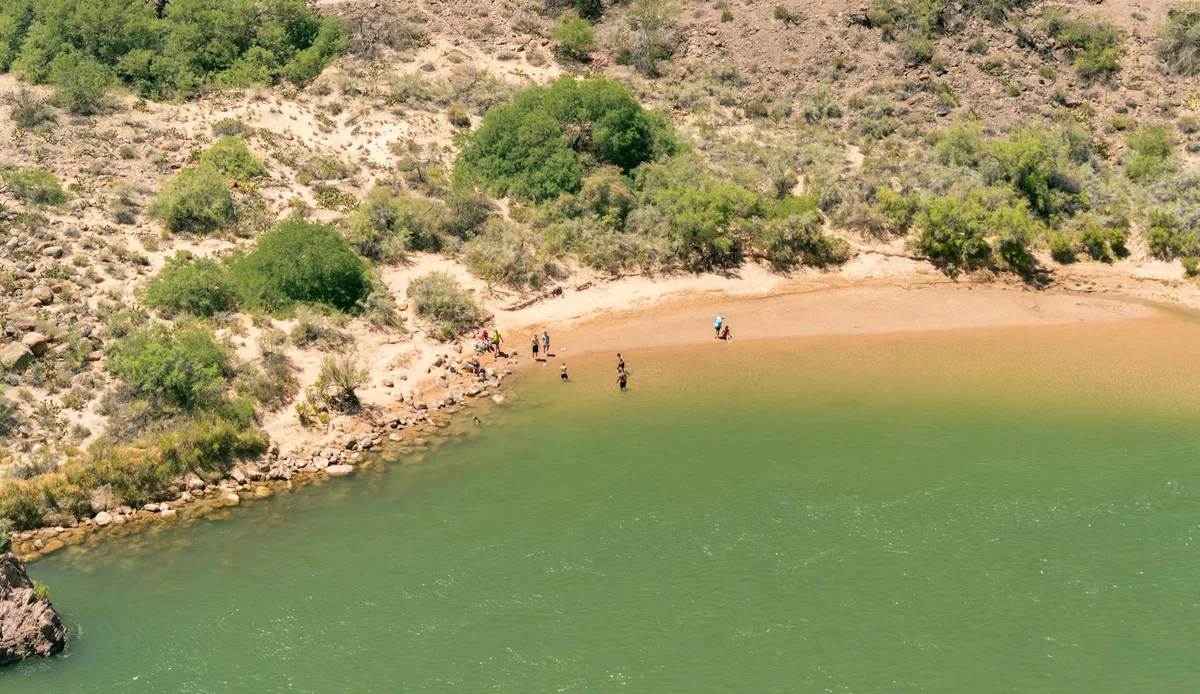 A wide aerial view shows a quiet green lake bordered by a sandy crescent shore and scrubby desert slopes, with a small group of people wading near the water’s edge. Sunlit tan hills and scattered shrubs frame the calm surface, giving the scene a warm late-day feel.