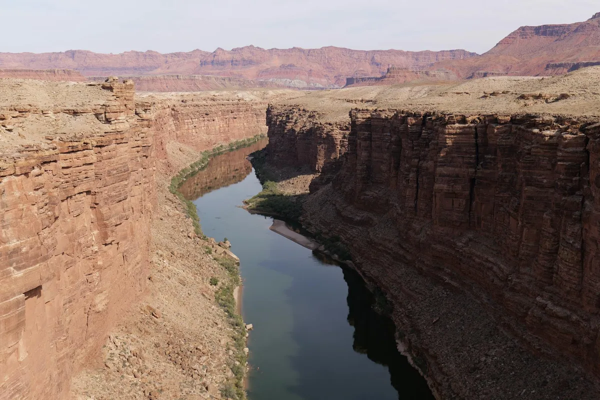 A narrow, calm river winds through steep red rock canyon walls, with the water reflecting the cliffs and distant mesas. Pale, scrubby slopes and layered rock textures frame the scene under soft golden light.