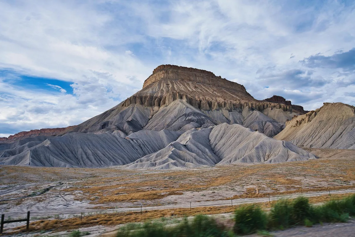 Towering gray and tan Book Cliffs rise in layered, eroded ridges beneath a wide blue sky with streaky clouds. A strip of dry grassland and a blurred fence line run across the foreground, giving the scene a broad, open feel.