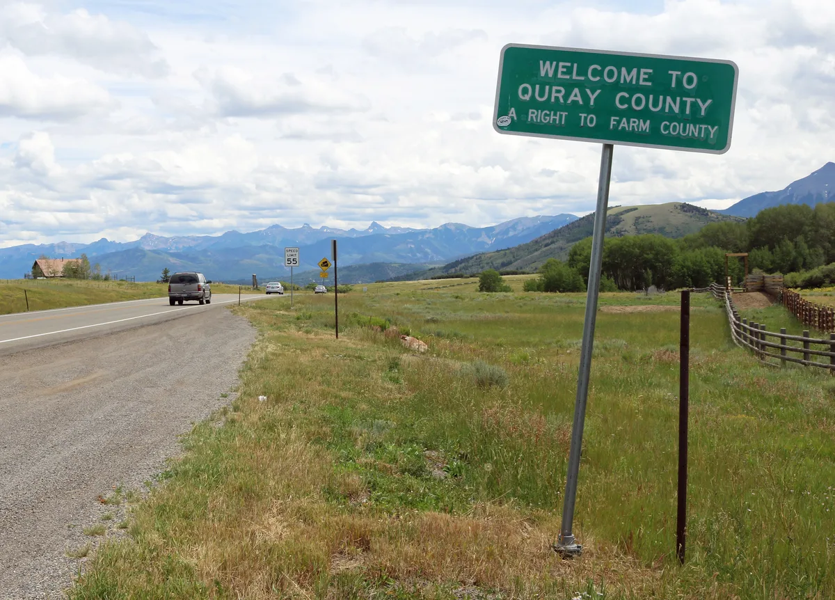 A roadside sign reading “Welcome to Ouray County” stands beside Highway 62, with a grassy shoulder, rail fence, and a few cars leading the eye toward the distant Wilson Range. Soft daylight and scattered clouds brighten the open meadow and layered blue mountains beyond.