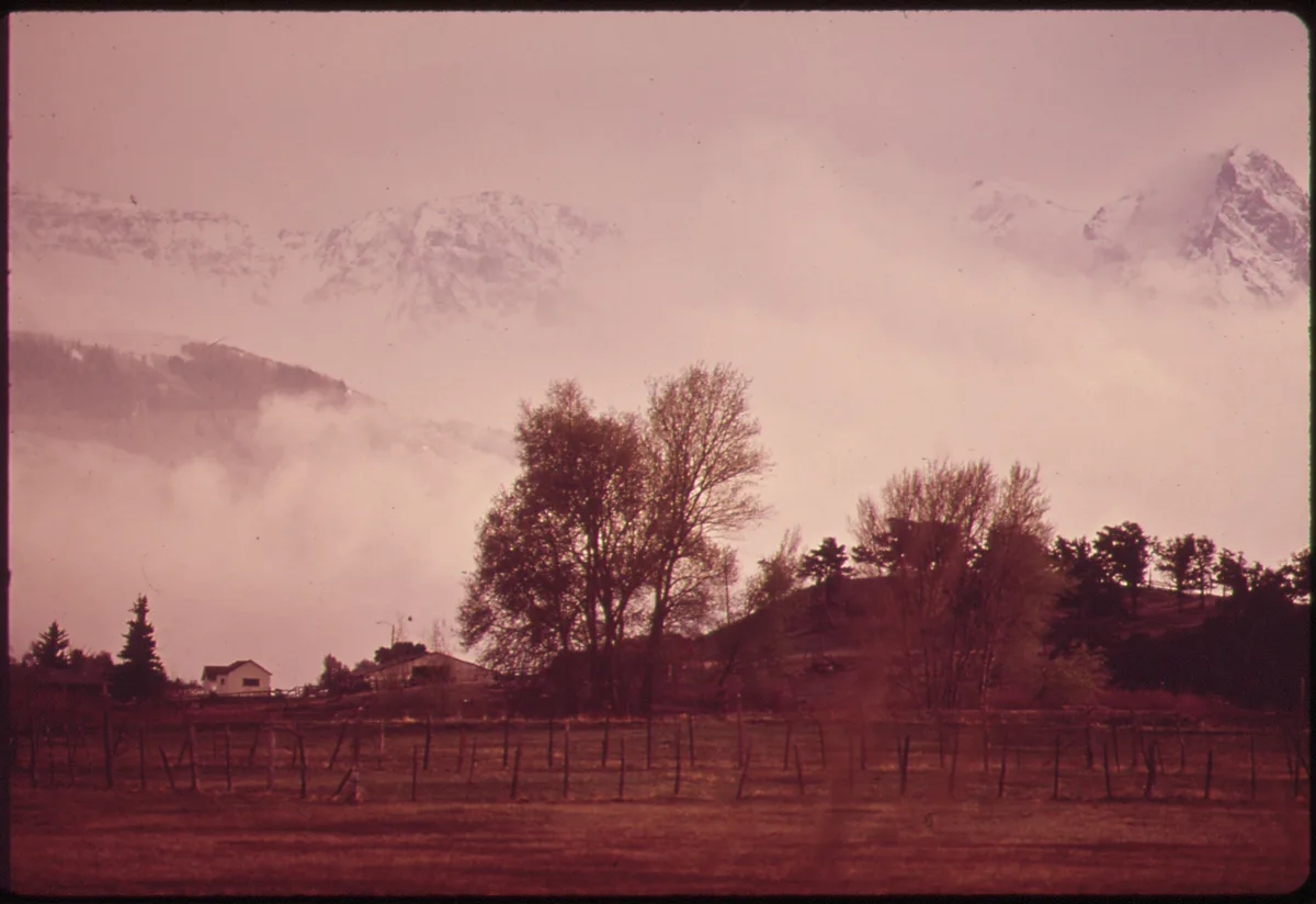 Low clouds hang over the Wilson Range at golden hour, with snow-dusted peaks fading into a pink haze above a dark foreground of meadow, bare trees, and a simple rail fence. A small house and rolling hillside sit beneath the mountain backdrop, giving the wide view a quiet Western feel.