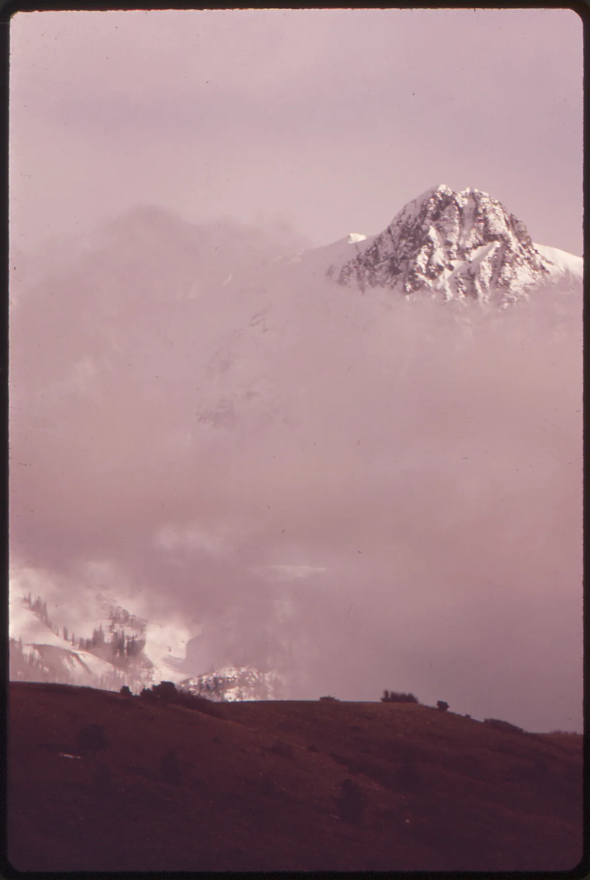 A snow-covered jagged peak rises above thick pink-tinted clouds, with only the upper mountain catching light while the lower slopes fade into haze. Dark rolling foreground hills anchor the bottom edge, creating a stark, layered alpine landscape.