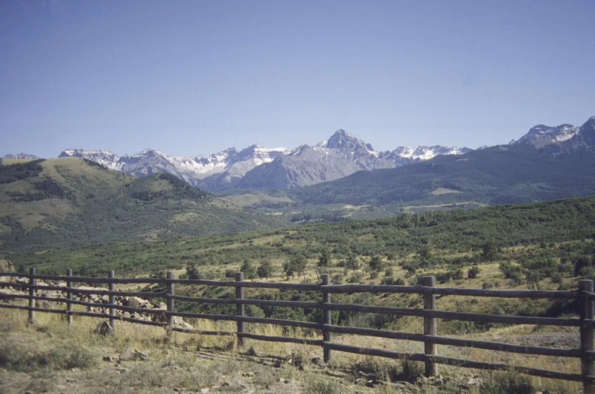 A wide view from Dallas Divide shows a weathered wooden rail fence crossing the foreground above rolling green meadows, with the jagged Wilson Range rising in the distance under a clear blue sky. Snow clings to the high peaks, and the late-day light gives the scene a crisp, open feel.