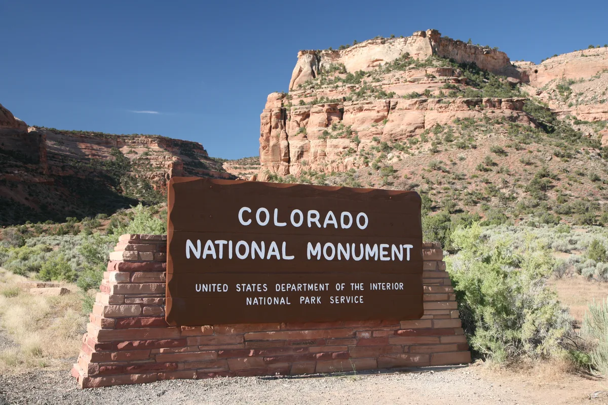 The Colorado National Monument entrance sign stands in the foreground, framed by sunlit red sandstone cliffs and scrub-covered slopes under a clear blue sky. The wide view emphasizes the layered rock formations and dry desert landscape behind the sign.