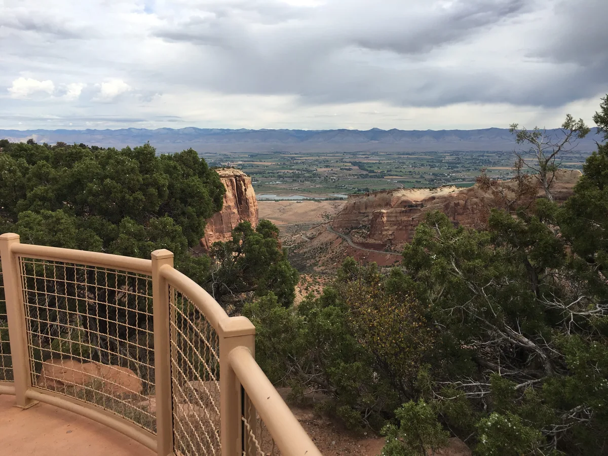 A tan lookout railing frames a sweeping view over red sandstone cliffs and a broad valley, with the Colorado National Monument’s layered mesas and distant mountains beneath a cloudy sky. Dense green shrubs and trees fill the foreground, contrasting with the warm rock and muted morning light.