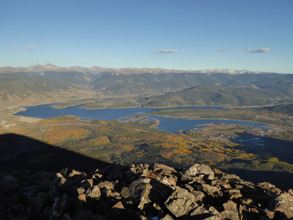 Wide view from a rocky overlook above Dillon Reservoir, with the blue water stretching across the valley below and snow-dusted mountain ridges lining the horizon. Golden light picks out patches of autumn color on the hillsides, while a dark foreground shadow frames the scene.