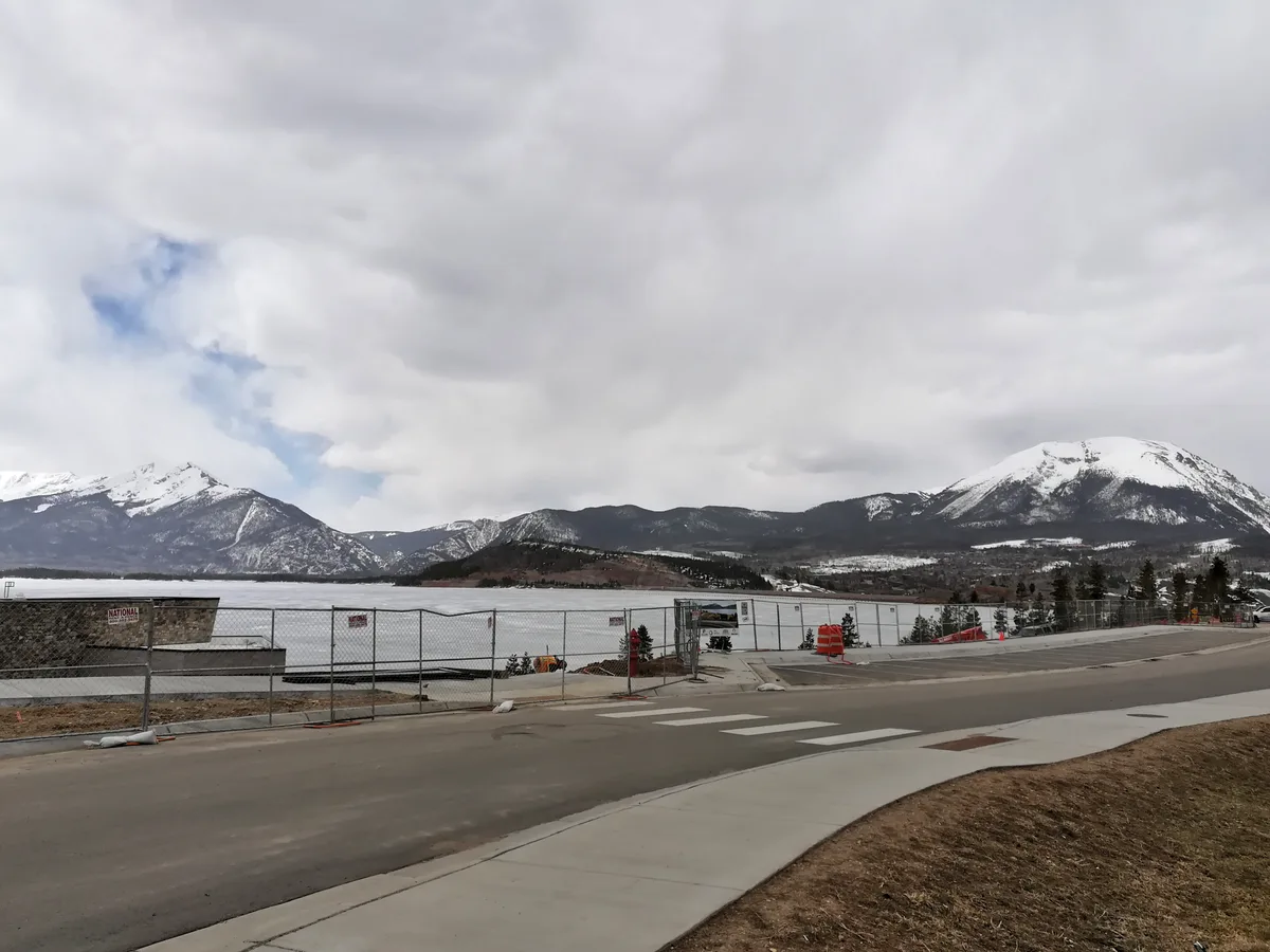 A wide view over Dillon Reservoir shows snowy mountain peaks under a thick, overcast sky, with the lake stretching across the middle distance. In the foreground, a paved road, sidewalk, and chain-link construction fence lead toward the shoreline, adding a developed edge to the alpine scene.