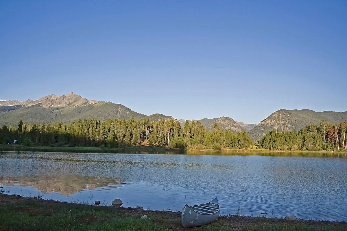 Golden light washes over Dillon Reservoir, with calm water reflecting the forested shoreline and jagged mountain peaks beyond. A lone canoe rests on the grassy bank in the foreground under a clear blue sky.