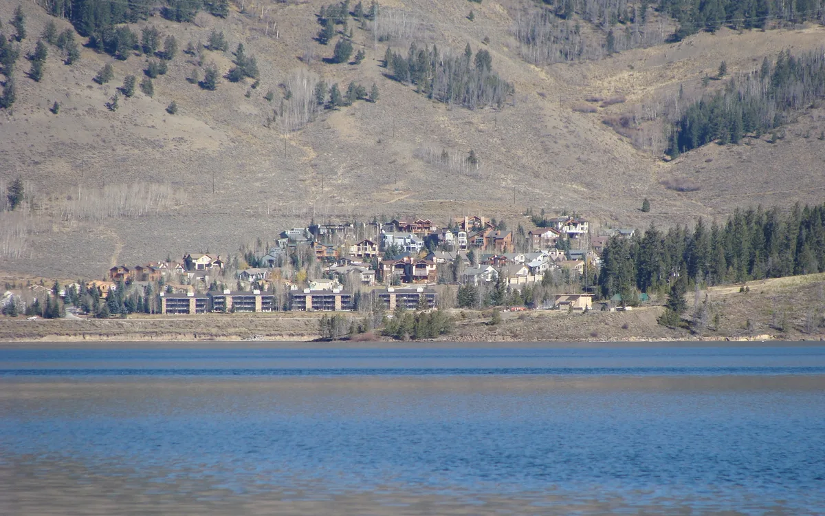 Blue Dillon Reservoir fills the foreground with a calm, lightly rippled surface, while a cluster of lakeside buildings and evergreen trees sits at the base of dry tan mountains beyond. The scene is framed in soft golden light, with the shoreline and hillside reflected faintly in the water.