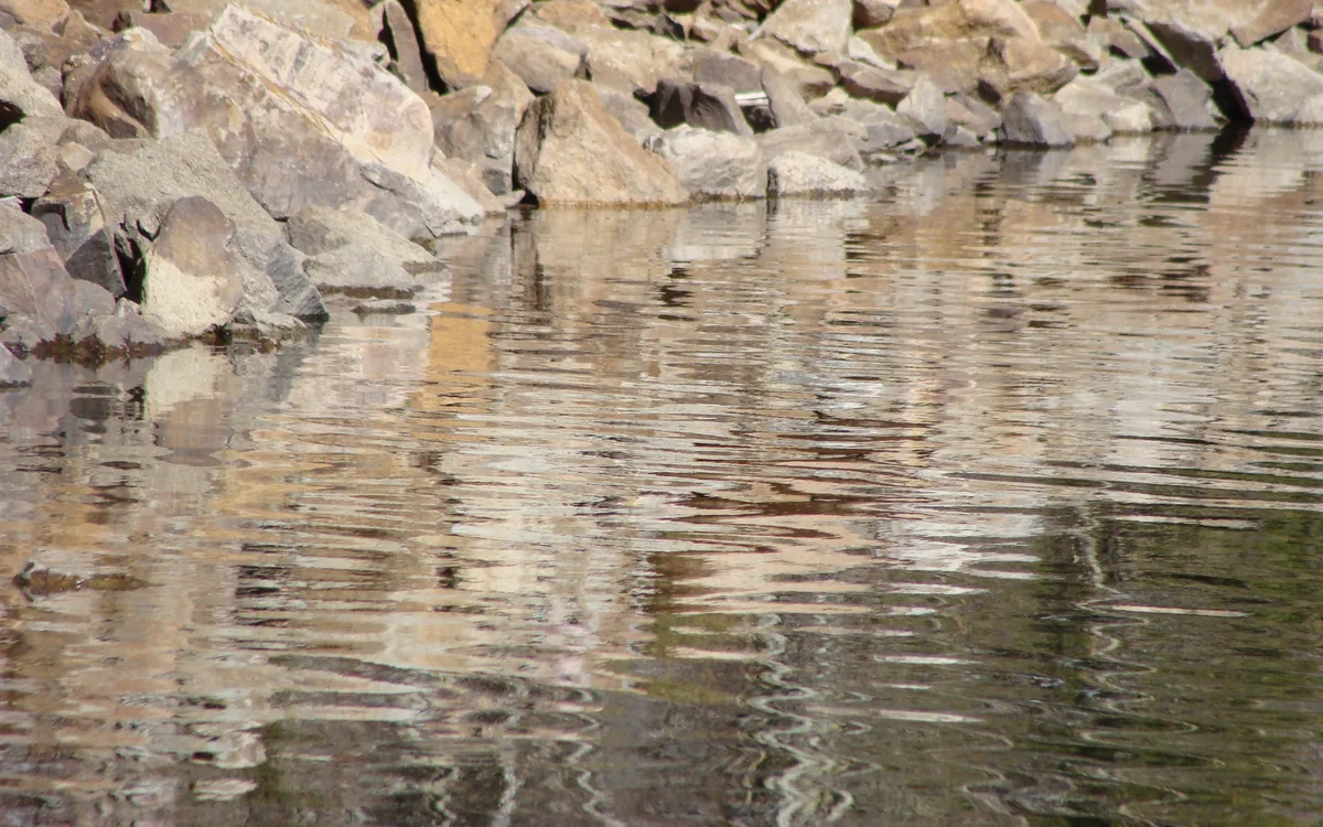 Golden light ripples across the reservoir’s surface, creating a shimmering reflection of the rocky shoreline above. The water fills most of the frame, with tan and gray boulders lining the top edge.