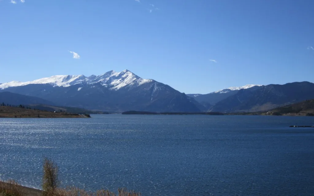 Calm blue Dillon Reservoir fills the foreground, with snow-covered mountain peaks and dark ridgelines stretching across the horizon under a clear sky. Sunlight glints off the water, while a narrow shoreline and a few small islands break up the wide alpine view.