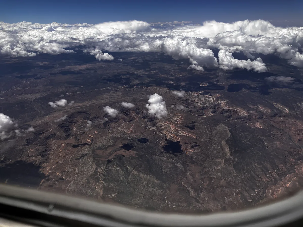 High-angle aerial view over a dark, rugged river canyon landscape, with pale winding rock layers and a few scattered clouds casting shadows below. The airplane window frame curves along the bottom edge, and a thick bank of bright white clouds fills the horizon under a deep blue sky.
