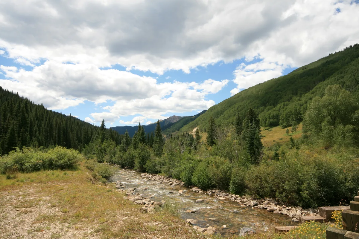 A clear mountain river winds through a green valley lined with dense evergreens and shrubs, with rounded slopes rising on both sides under a sky of thick white clouds and blue openings. The rocky streambed and sunlit grasses give the scene a late-summer feel.