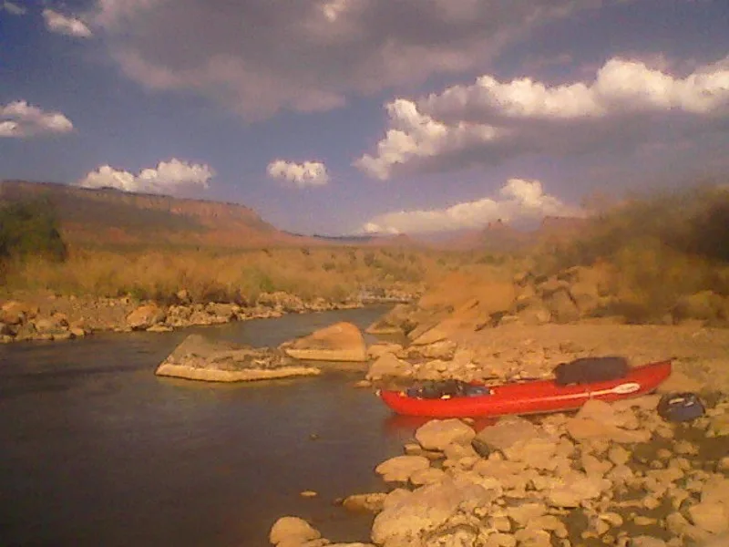 A red canoe rests on a rocky riverbank beside the low, dark water of the Dolores River, with red sandstone boulders and cottonwood-lined canyon slopes stretching into the distance. Soft golden-hour light warms the scene under a sky filled with scattered white clouds.