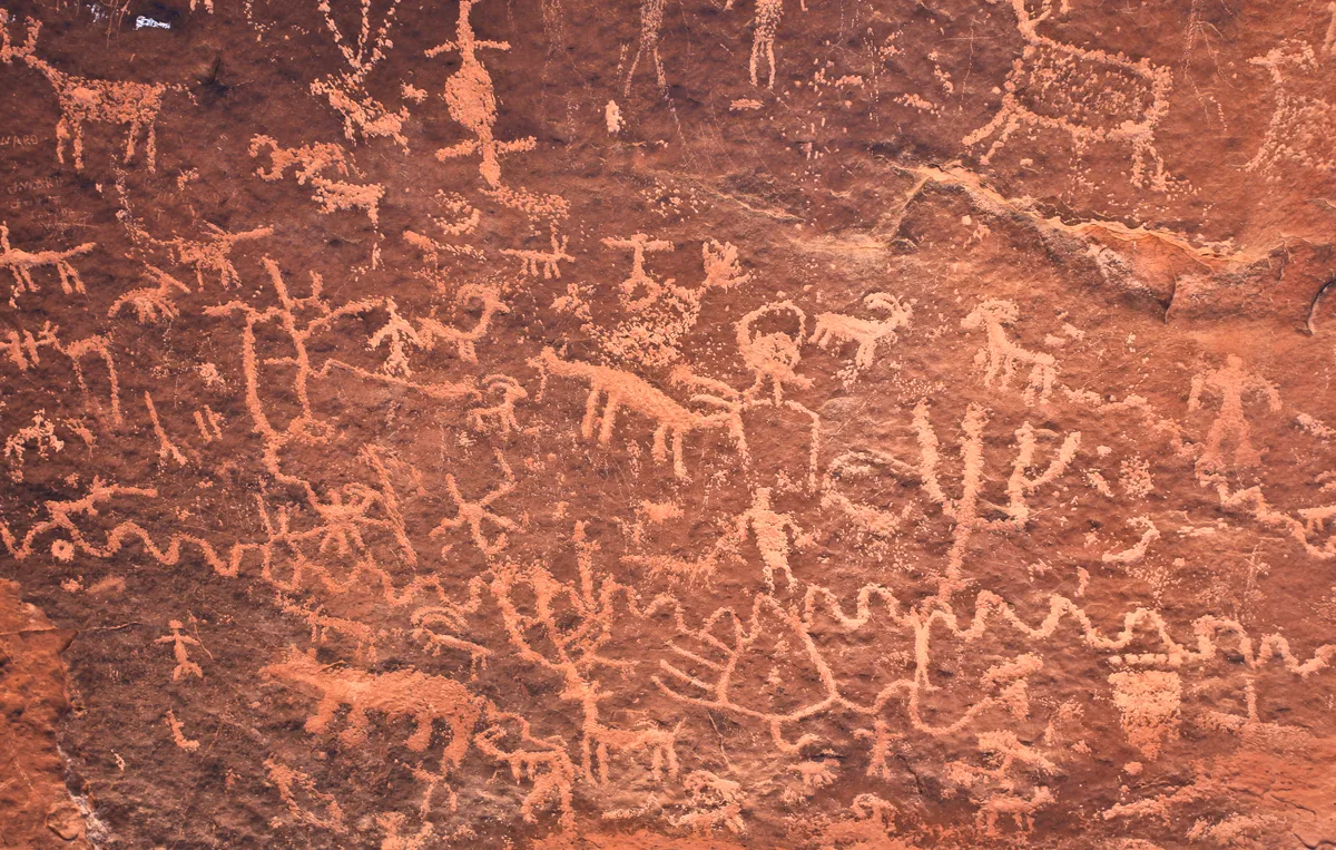 Close-up of a reddish sandstone wall covered with pale Ancestral Puebloan petroglyphs, including human figures, animals, and abstract symbols. Warm morning light brings out the rough texture and layered desert varnish of the rock.