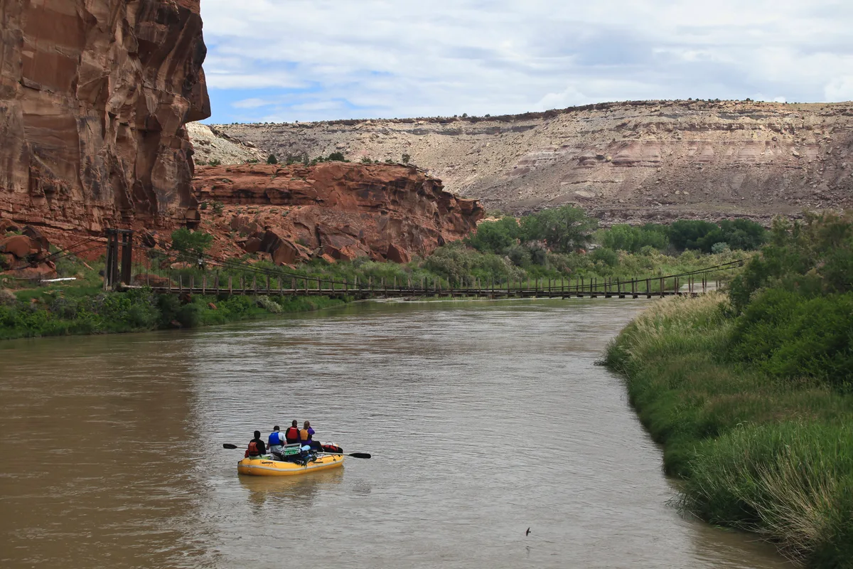 A yellow raft with four paddlers floats down a muddy river through a red sandstone canyon, with a narrow suspension bridge stretching across the water in the middle distance. Layered cliffs and pale desert hills rise under a cloudy sky, with green shrubs lining the riverbanks.