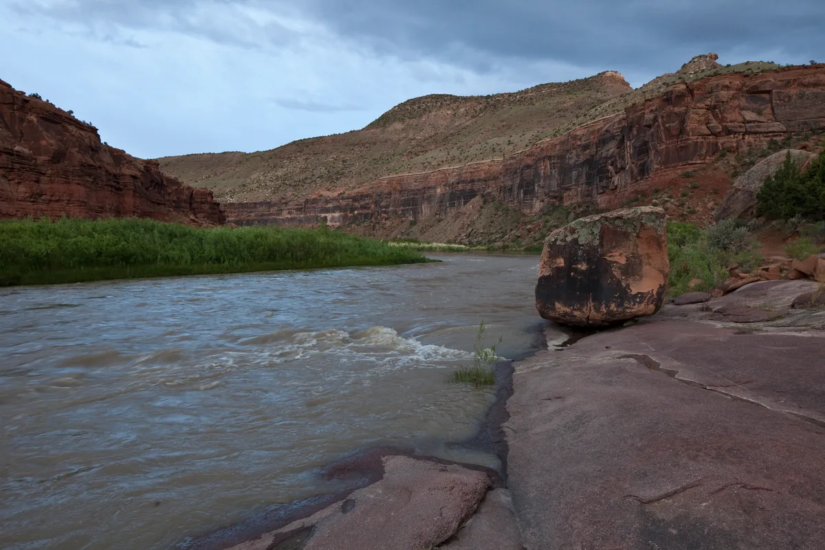 A broad river curves through a red sandstone canyon, with dark striped cliffs rising on both sides under a cloudy morning sky. A large boulder sits on the rocky bank in the foreground, while green brush lines the water’s edge.