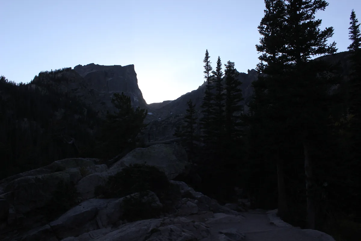 Dark pine silhouettes and boulders frame Dream Lake at dawn, with Hallett Peak and the surrounding cliffs forming a jagged horizon against a pale blue sky. The low light leaves the foreground in deep shadow, emphasizing the dramatic mountain backdrop.