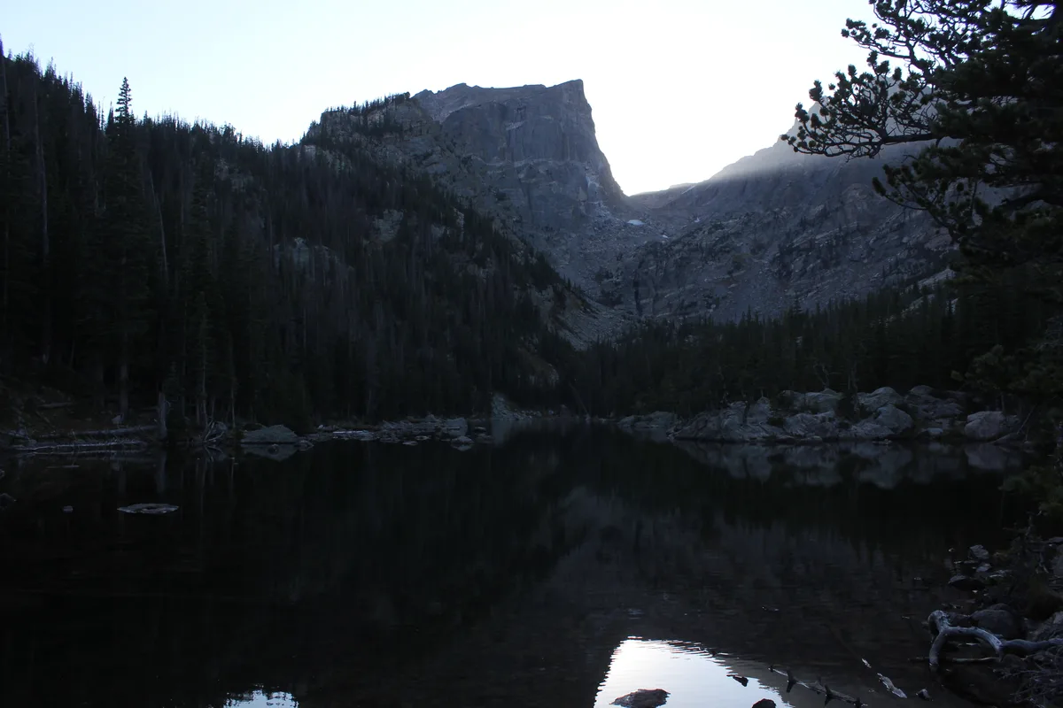 Dark, still Dream Lake reflects the surrounding pines and the steep gray face of Hallett Peak, while the bright sky glows behind the ridge. A few boulders and a twisted tree branch frame the foreground, giving the scene a quiet, early-light feel.