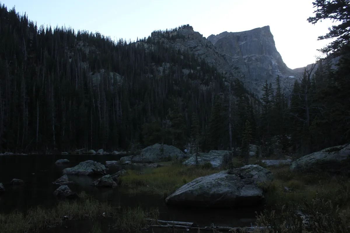 Dark, still water and scattered boulders fill the foreground, with dense pine forest climbing the slopes behind Dream Lake. Hallett Peak rises sharply in the background, silhouetted against a pale sky at dusk.