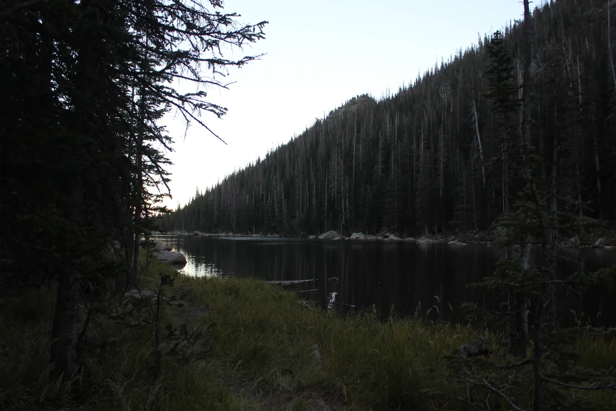 A dark, glassy alpine lake sits between steep, pine-covered slopes, with the pale sky reflected in the still water. Tall evergreens frame the foreground, and the scene feels quiet and subdued at dusk.