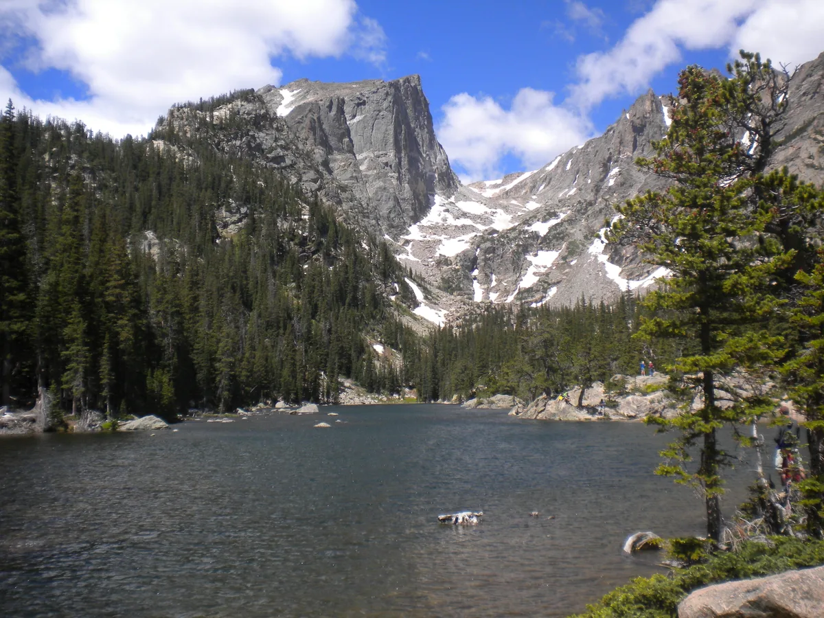 A wide view across Dream Lake shows dark water in the foreground, dense evergreen forest on the left, and steep gray cliffs and snow patches rising to Hallett Peak under a bright blue sky with scattered clouds. A pine tree and a few hikers frame the right edge, emphasizing the alpine scale of the scene.
