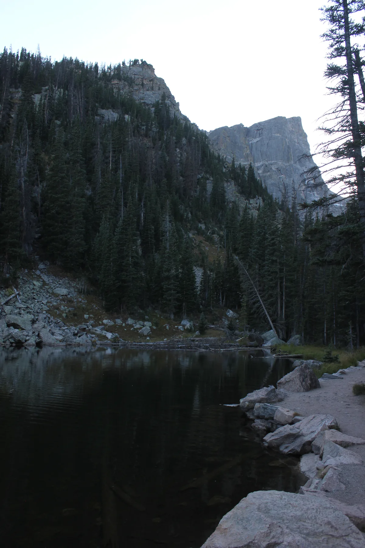 A dark alpine lake sits below steep pine-covered slopes and a sheer gray cliff, with its still surface reflecting the surrounding trees. Large rocks and a narrow trail edge the right side, while the bright sky silhouettes the rugged peaks above.