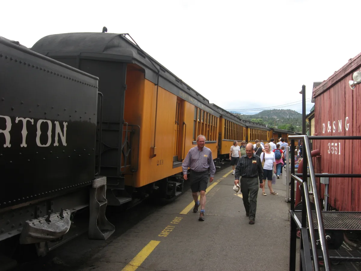 Passengers walk along the platform beside a row of Durango & Silverton Narrow Gauge Railroad cars, including a black car and a weathered red car marked “D&SNG 363,” under a bright overcast sky. The train stretches into the distance with green hills and power lines visible beyond the station.