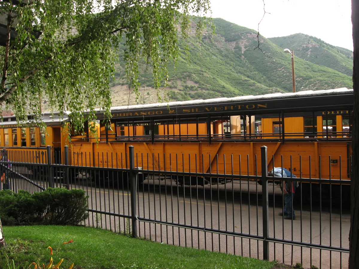 Orange Durango & Silverton passenger cars sit on the tracks behind a black metal fence, framed by green grass and hanging tree branches in the foreground. Green, shrub-covered mountains rise in the background under a bright morning sky.