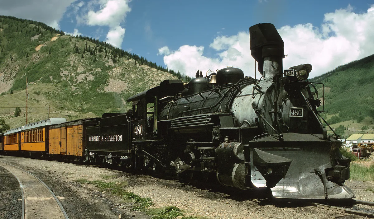 A black steam locomotive numbered 480 pulls orange passenger cars along a curving narrow-gauge track, with sunlit green mountain slopes rising behind it. Wisps of steam and the coal-fired engine details stand out against the bright blue sky and scattered clouds.