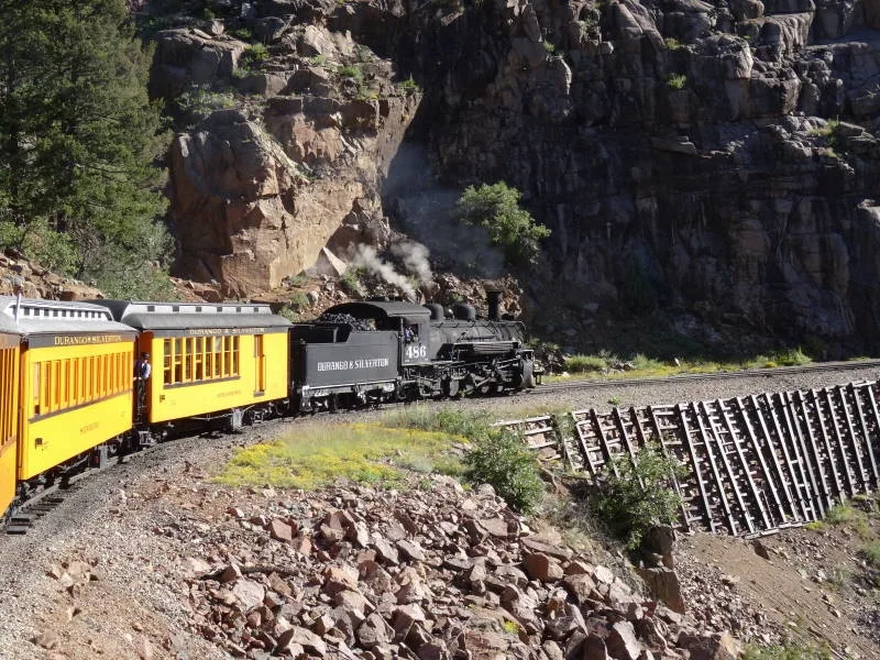 A black steam locomotive pulls bright yellow Durango & Silverton passenger cars along a narrow track cut into a rocky canyon wall, with smoke drifting against the dark cliff. The morning light highlights the steep ledge, gravel slope, and timbered retaining wall beside the curve.