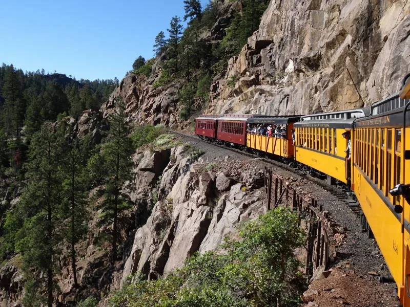 A narrow-gauge steam train curves along a cliffside ledge above a forested canyon, with bright yellow and red cars hugging the rock wall. Clear morning light highlights the rugged granite slopes and the steep drop-off beside the tracks.
