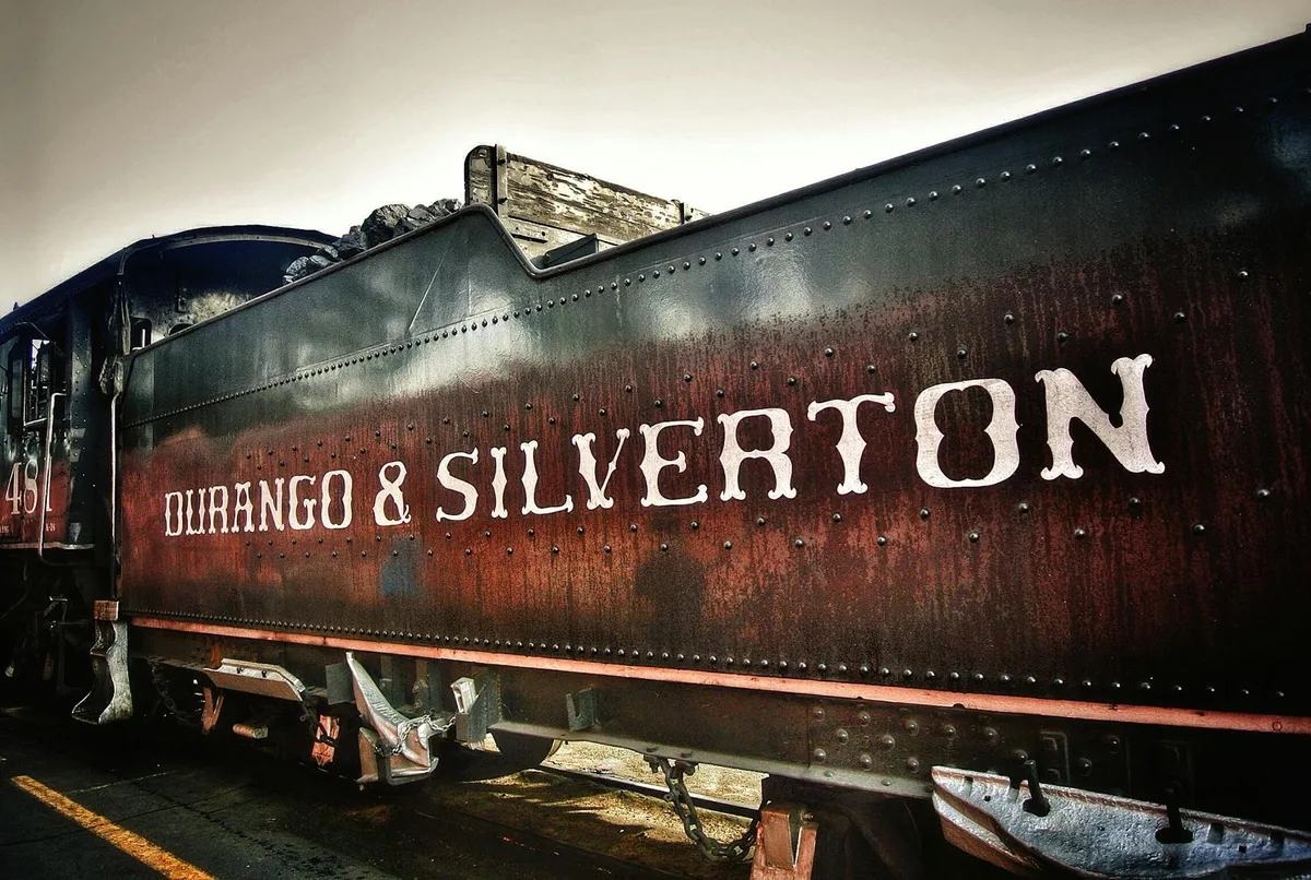 Close-up of a weathered black steam locomotive tender with riveted metal panels and large white “Durango & Silverton” lettering, lit by soft morning light. The low angle emphasizes the train’s heavy machinery, with coal piled above the tender and a hint of the engine cab at left.