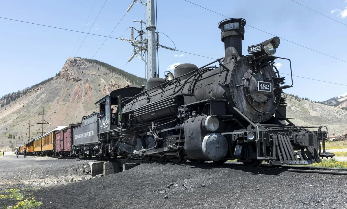 A black steam locomotive numbered 482 leads a narrow-gauge train past the base of a rocky mountain under a clear blue sky, with yellow passenger cars trailing behind. Power lines and a gravel turnout frame the scene in bright morning light.