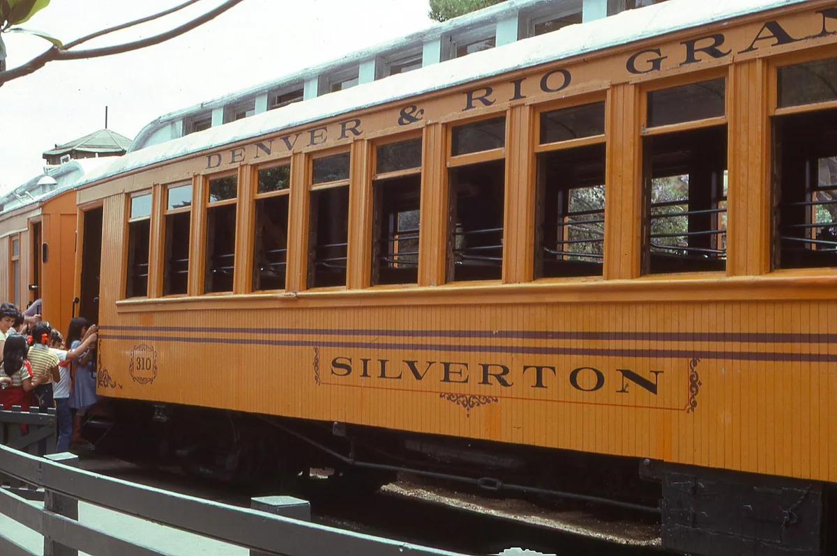 A mustard-yellow historic railcar with “Denver & Rio Grande” and “Silverton” painted across the side fills the frame, its open windows and green roof details visible in soft morning light. On the left, a small group of passengers gathers beside the train at the platform.
