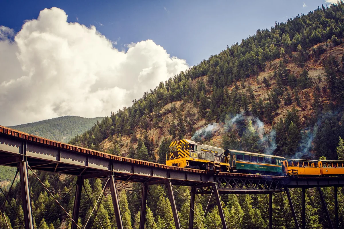 A coal-fired narrow-gauge steam train crosses a tall steel trestle clinging to a steep, pine-covered canyon wall, with a puff of smoke trailing behind the engine. Bright morning light and towering white clouds frame the mountain landscape below.