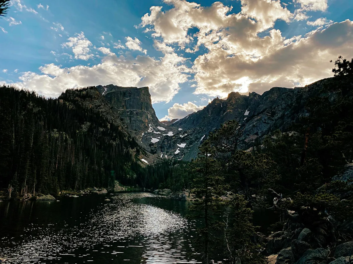 Dark green Emerald Lake sits in a rocky alpine basin, with steep granite walls and lingering snow patches rising behind it. Low morning sun breaks through dramatic clouds, brightening the water’s rippled reflection and the silhouetted evergreens along the shore.
