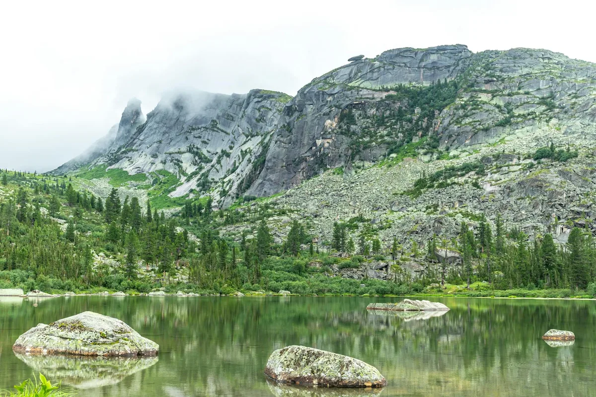 Still green alpine lake water fills the foreground, dotted with partially submerged boulders that reflect in the glassy surface. Steep gray cliffs and pine-covered slopes rise behind it, with low clouds clinging to the mountain peaks in soft morning light.
