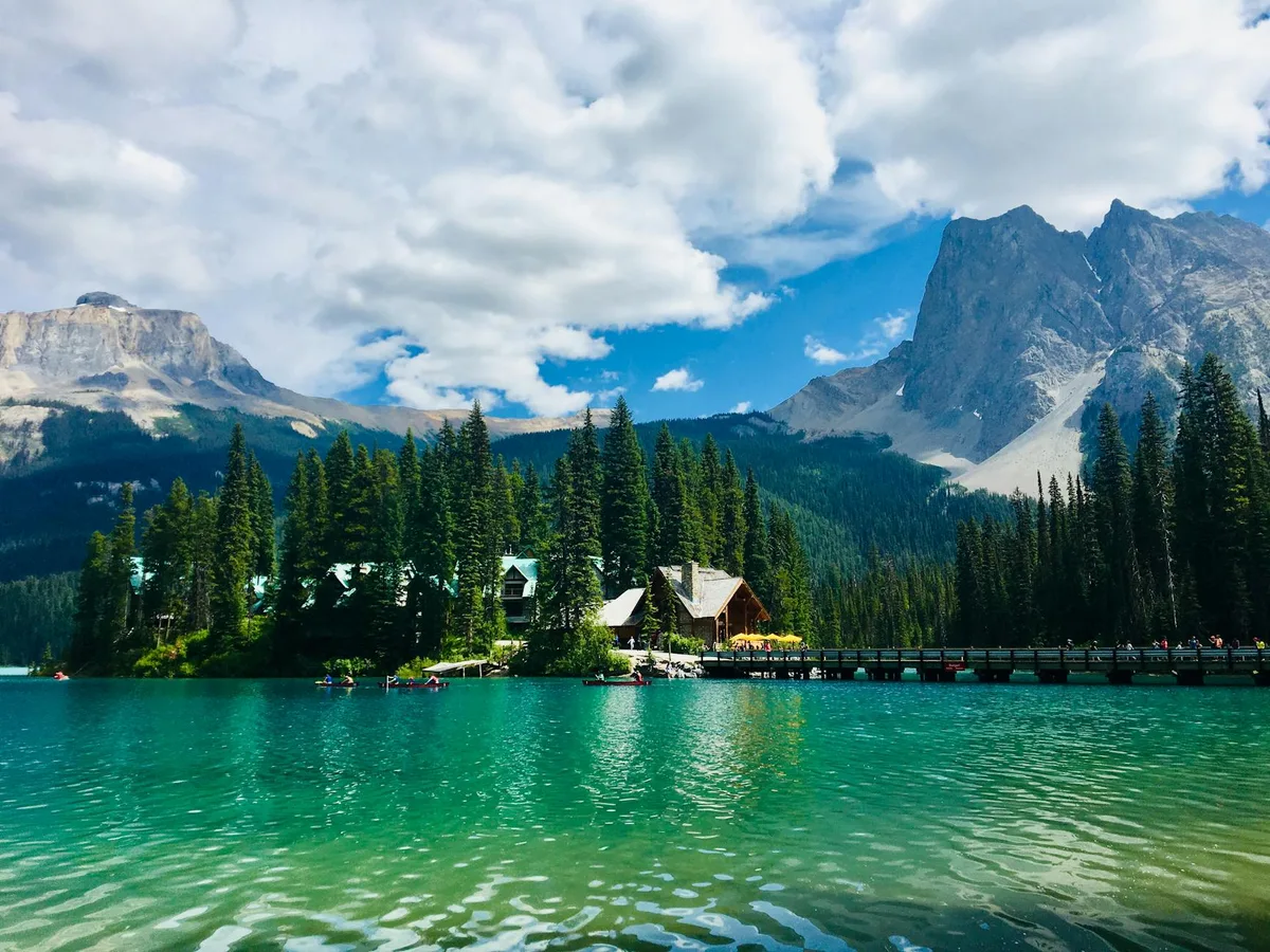 Turquoise Emerald Lake fills the foreground, rippling under a bright morning sky, with evergreens, a few lakeside buildings, and a long footbridge along the shore. Towering gray peaks rise behind the forest, their steep, rocky faces framed by drifting white clouds.