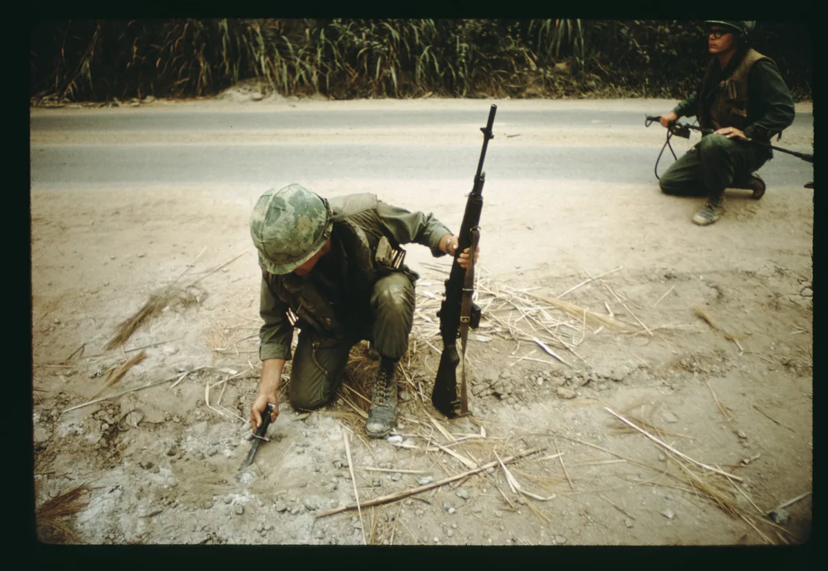Two soldiers in olive uniforms kneel on a dusty roadside, one probing the ground with a hand tool while holding a rifle upright. A second soldier watches from the road shoulder, with tall grass and a faded lane forming the background.
